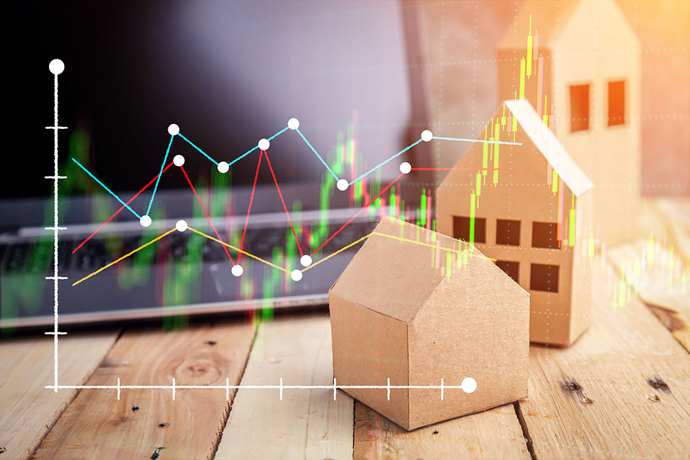 A red and green line chart overlaying three wooden models of different size houses and a laptop computer